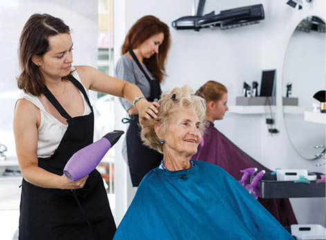 Professional female stylist making hairdo for senior woman in salon, using hair dryer 